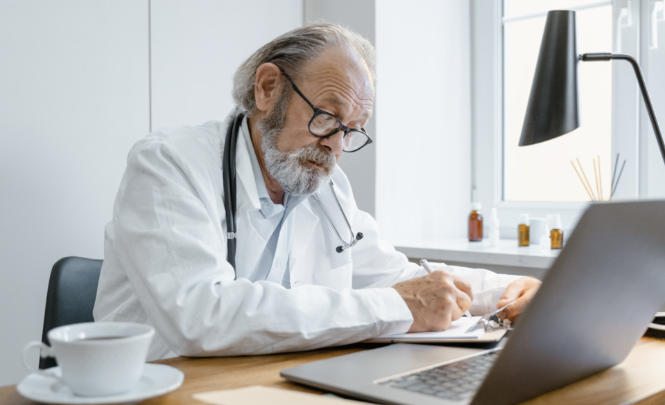 Médico de jaleco e estetoscópio fazendo anotações em sua mesa de trabalho